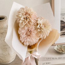 Load image into Gallery viewer, A bouquet of dried or pressed large pale peach and cream flowers wrapped in white tissue paper and a blush pink ribbon, styled on a tabletop with a ceramic vase, framed print, and books in the background.
